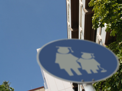 Straßenschild mit zwei sich an die Hand nehmenden Kindern mit Doktorhüten vor Unigebäude